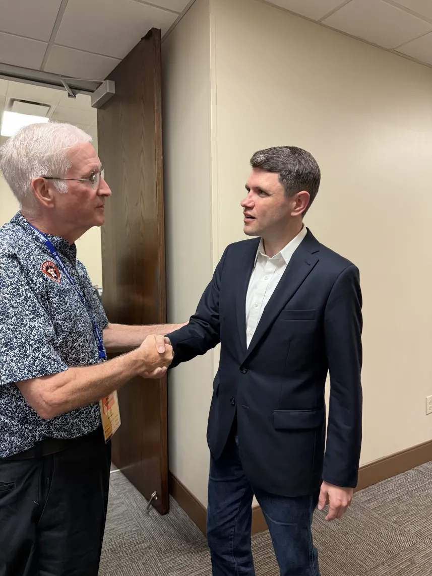 Two men shaking hands indoors, one in a suit and the other in casual shirt.